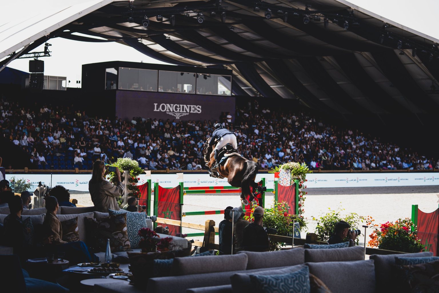 Show jumper clearing a fence before a full grandstand, seen from the VIP lounge